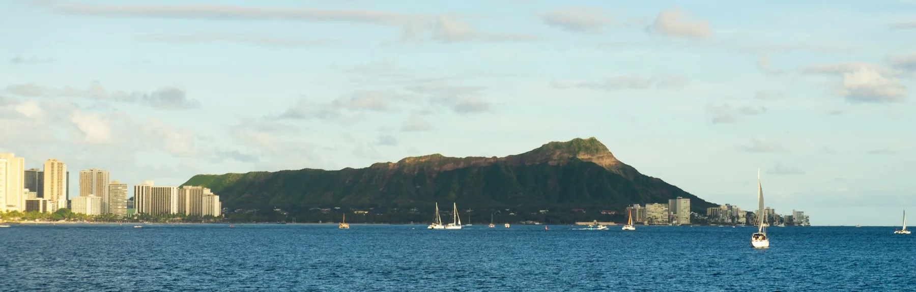 Der Diamond Head und die Skyline von Waikiki über blauem Wasser, in der Ferne Segelboote
