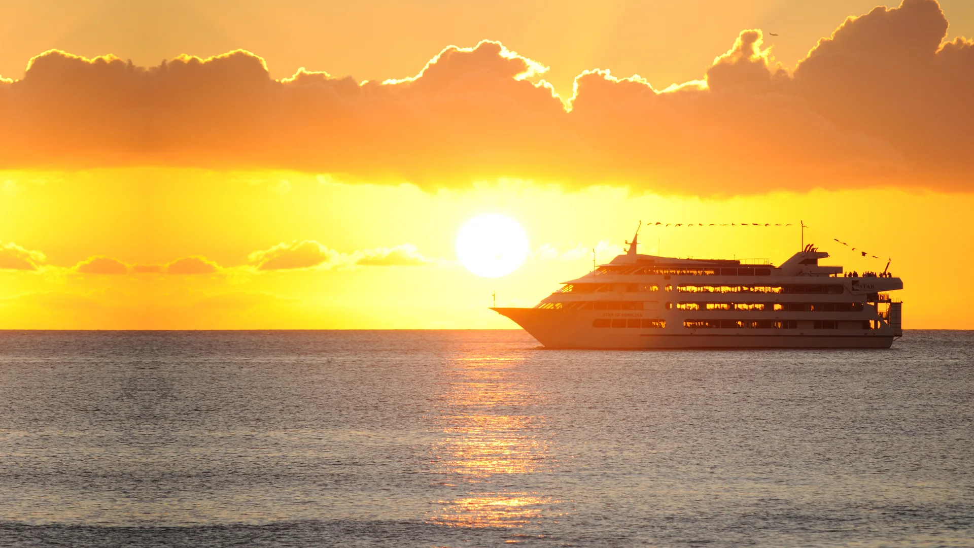 Die Star of Honolulu mit vier Decks auf Fahrt unter leuchtenden Wolken über ein goldenes Sonnenuntergangsmeer vor Honolulu