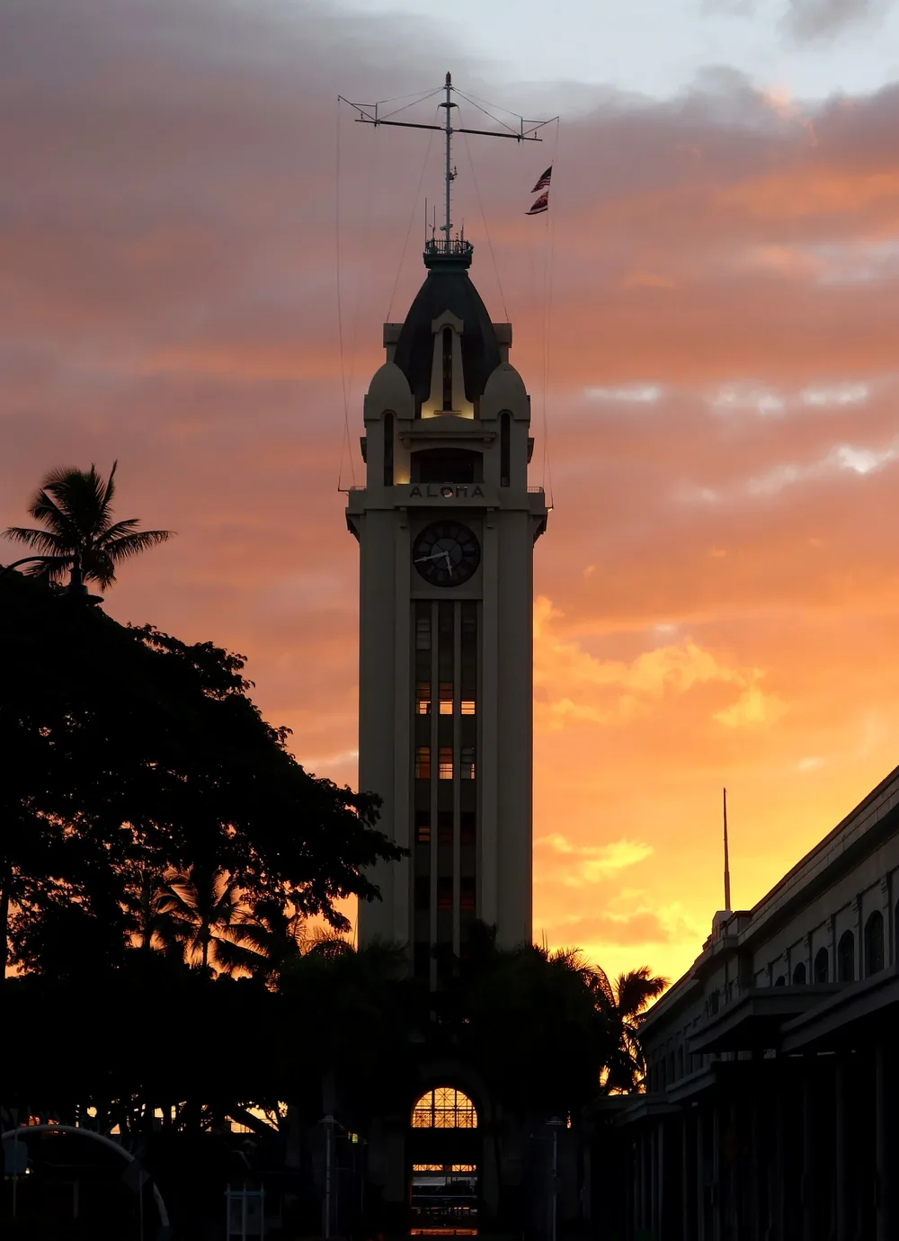 Der Aloha Tower als Silhouette vor einem orange-rosa Abendhimmel über Palmen